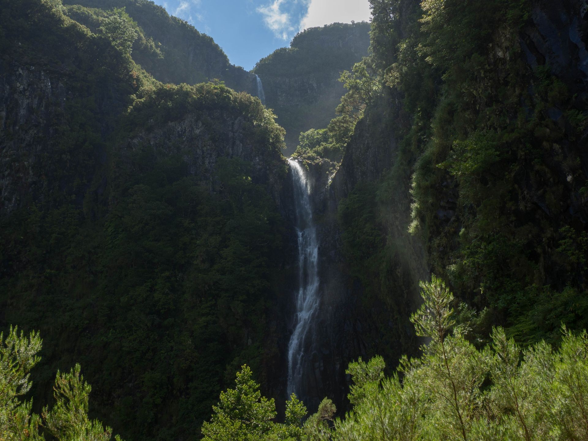 Tall waterfall cascading down a lush, green mountainside under a bright blue sky with scattered clouds.