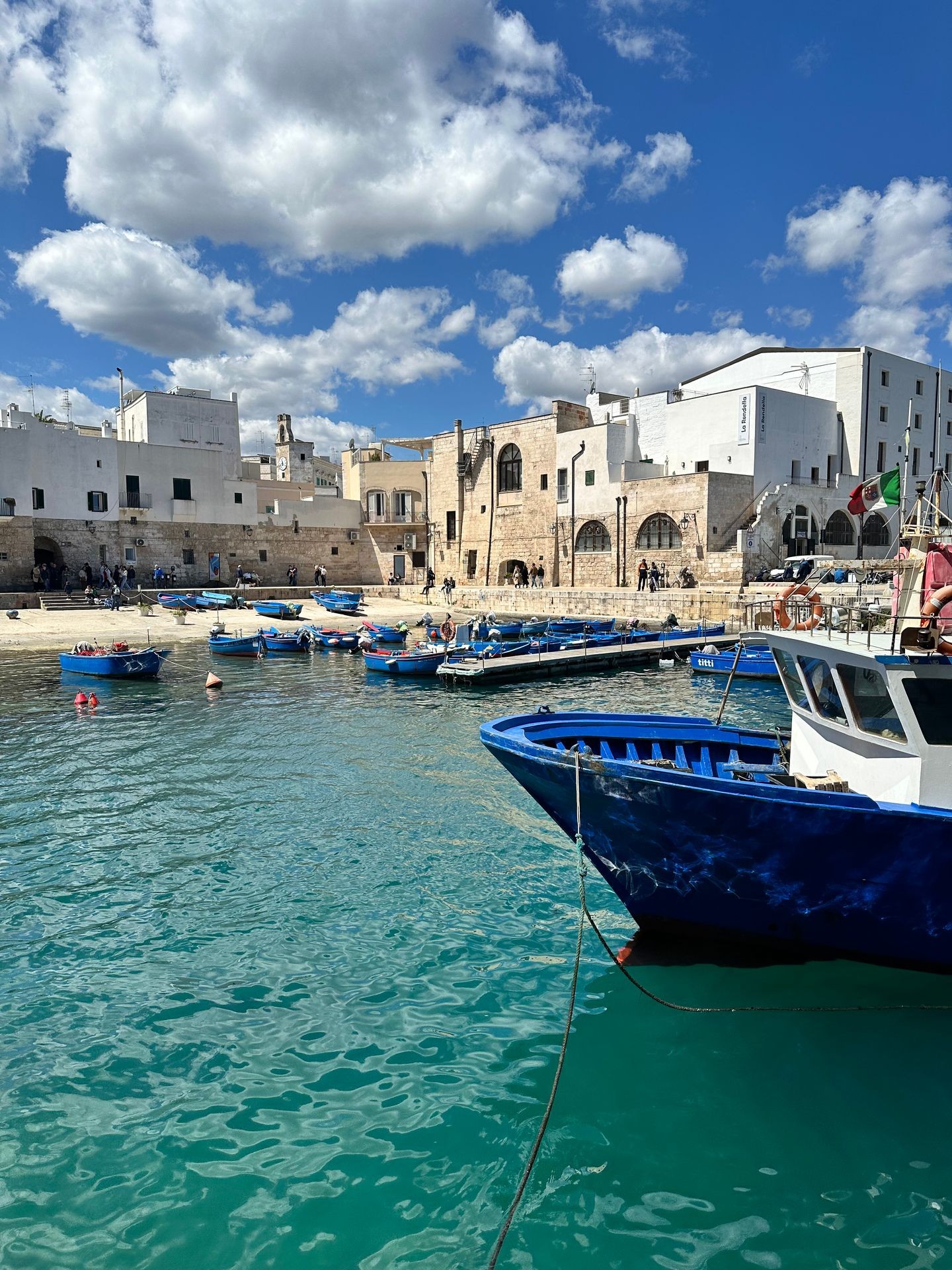 Seaside town with white buildings, blue boats in harbor, and cloudy sky.