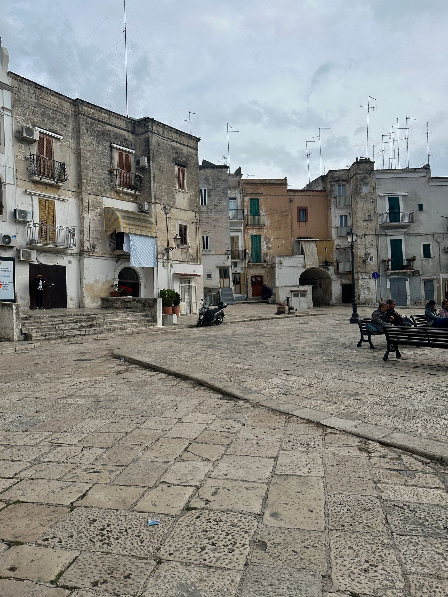 Old stone buildings with balconies in a European square, featuring benches and people sitting.