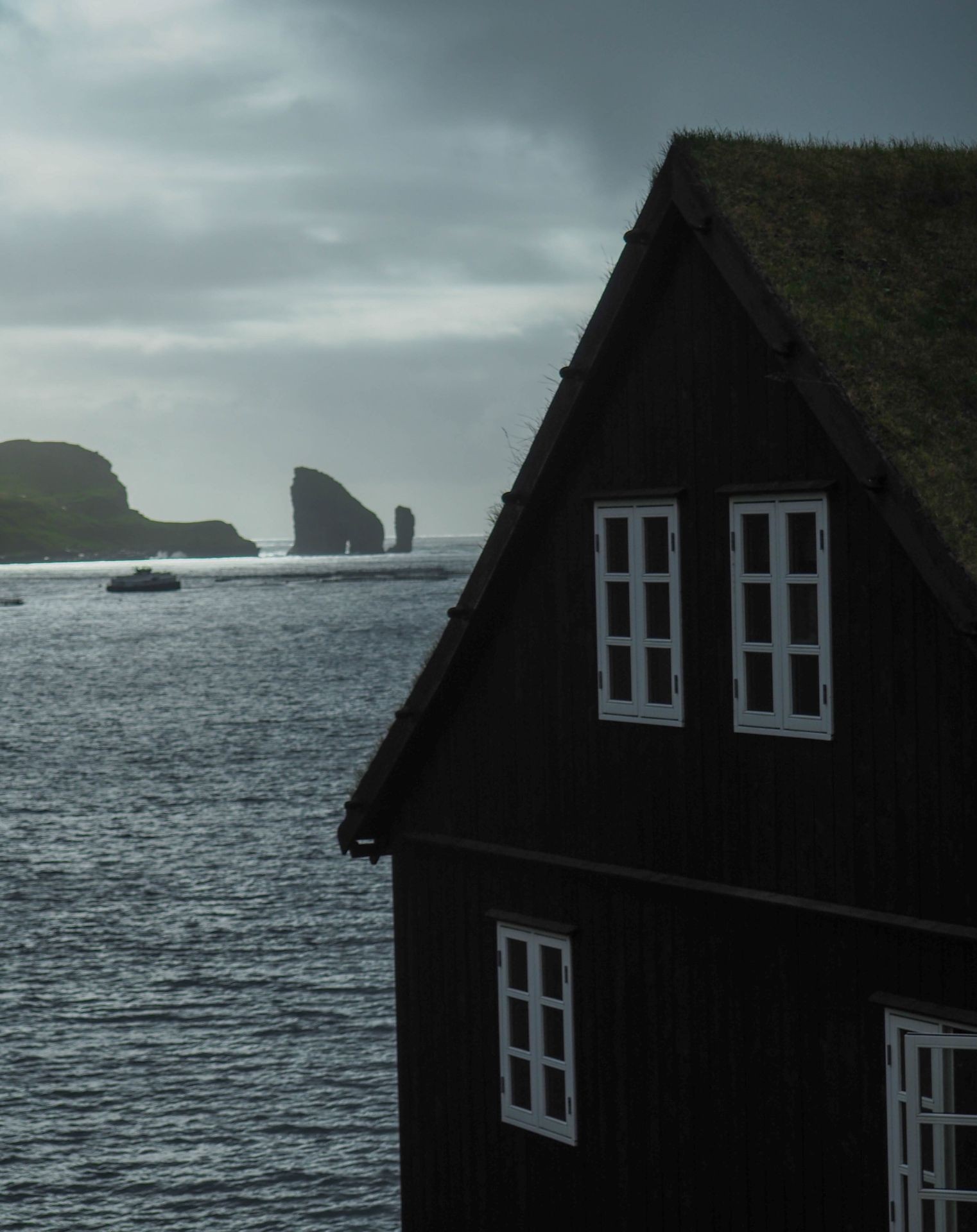 Coastal scene with a wooden house by the water, cloudy sky, and distant rocky cliffs.