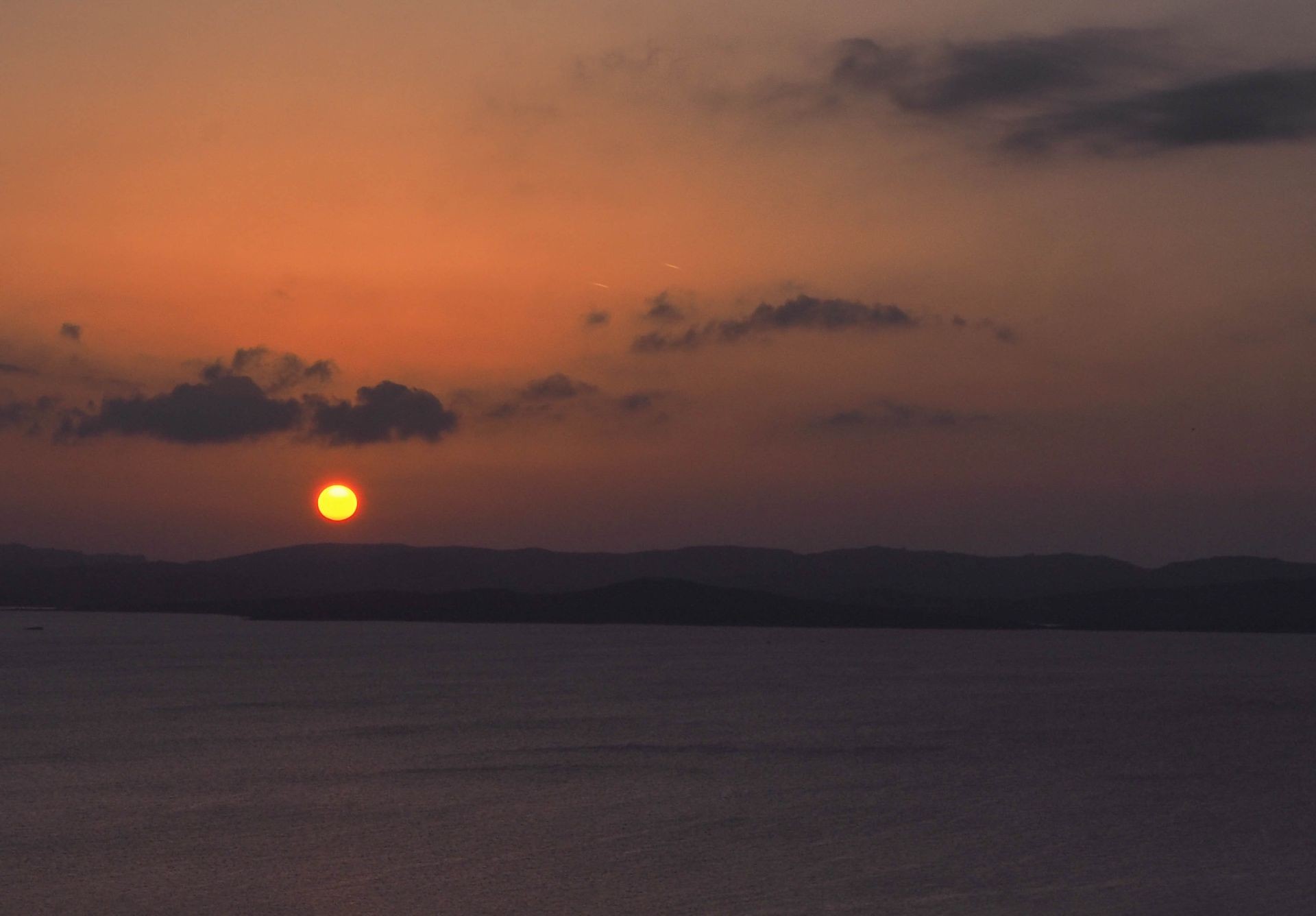 Sunset over calm sea with silhouetted hills and scattered clouds on the horizon.