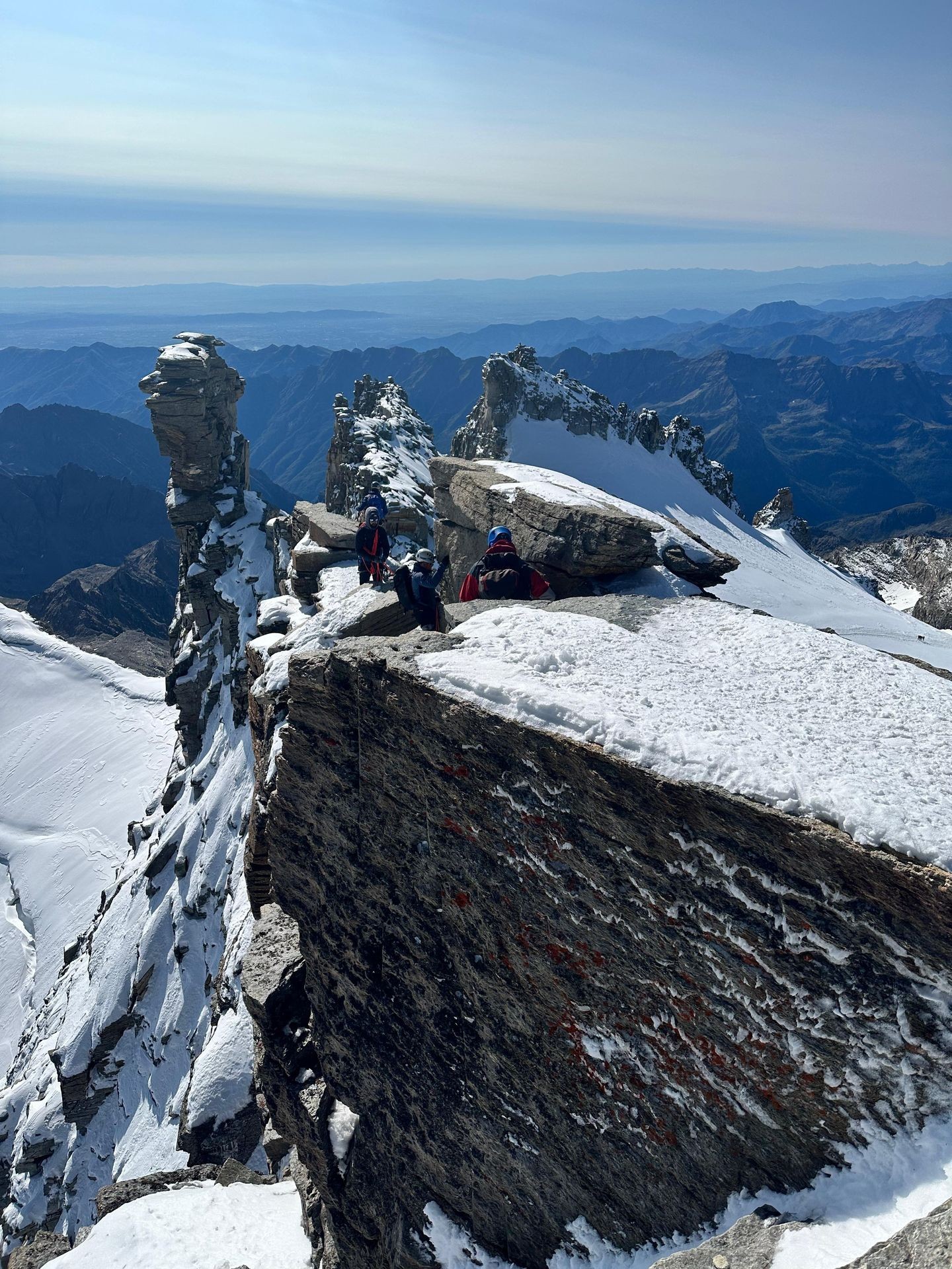 Mountaineers climbing a snowy ridge with rocky outcrops and distant mountains in the background.