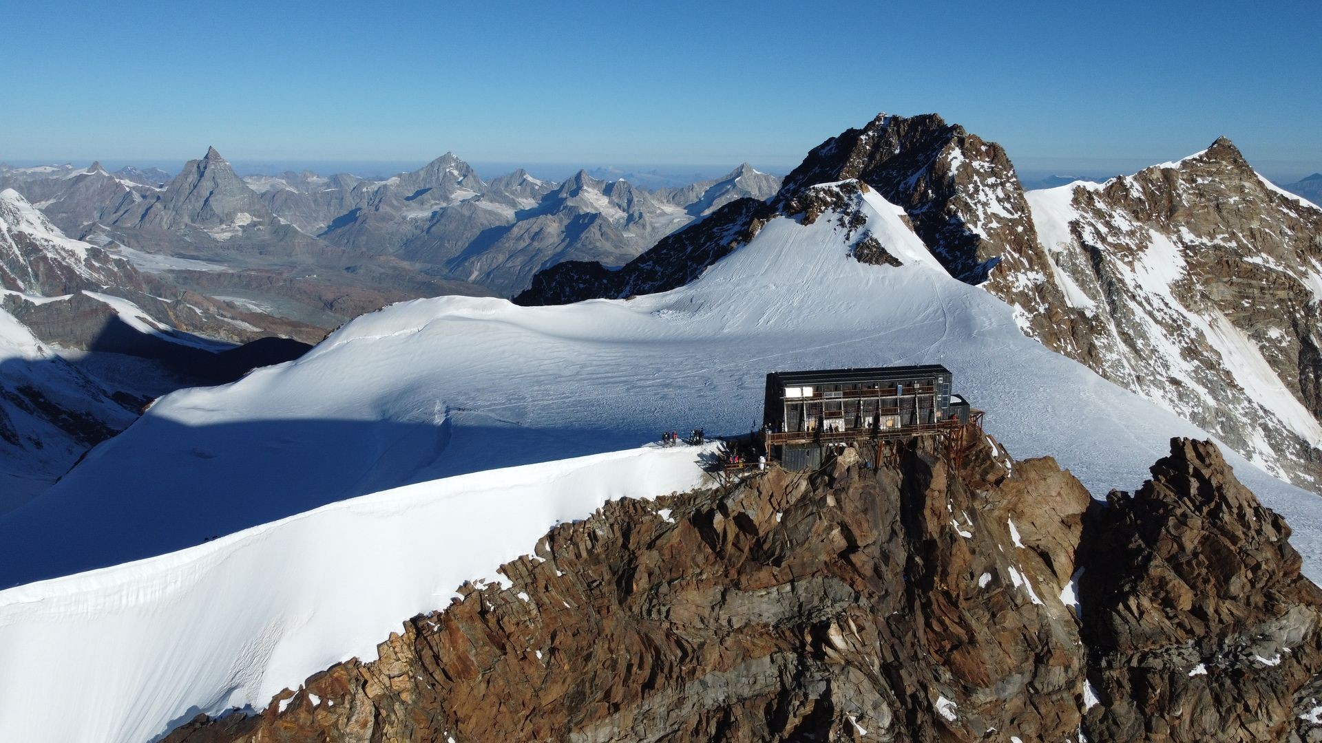 Mountain hut perched on a snowy ridge with rocky peaks in the background under a clear blue sky.